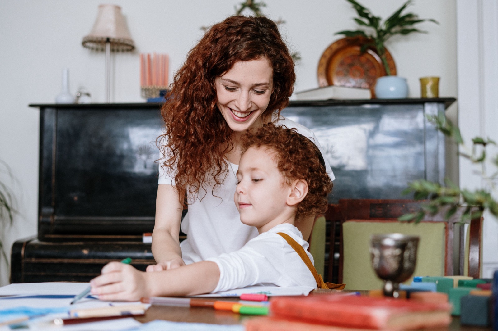 A mom leaning in with a big smile as her curly-haired son draws with markers at the table, an upright piano behind them.