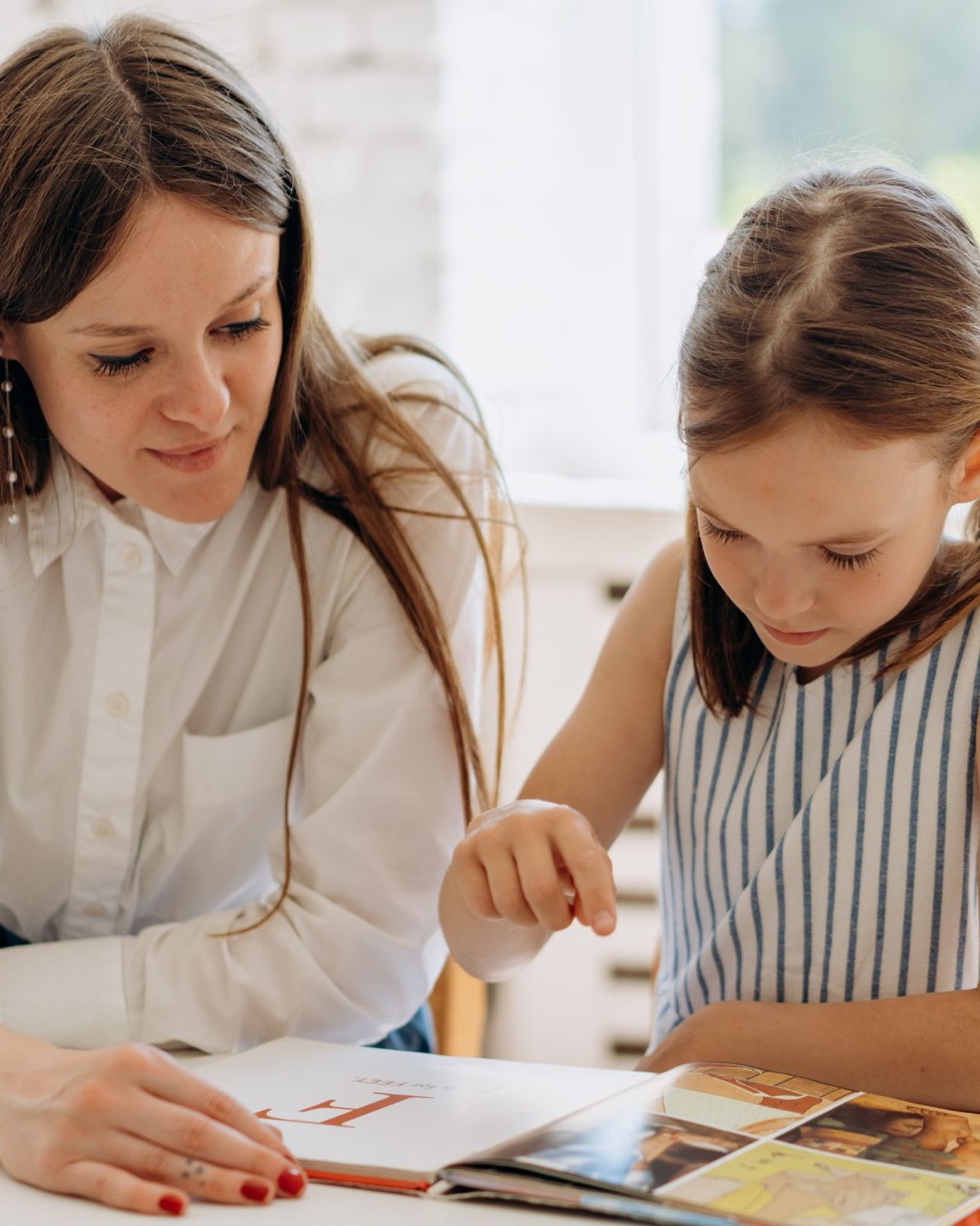 A mother sits beside her daughter at a table, guiding her through a book as the girl points to the page.